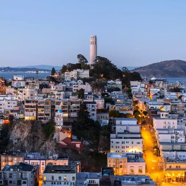 San Francisco's 屁股塔 at dusk, with lighted streets before it and the San Francisco Bay behind it.